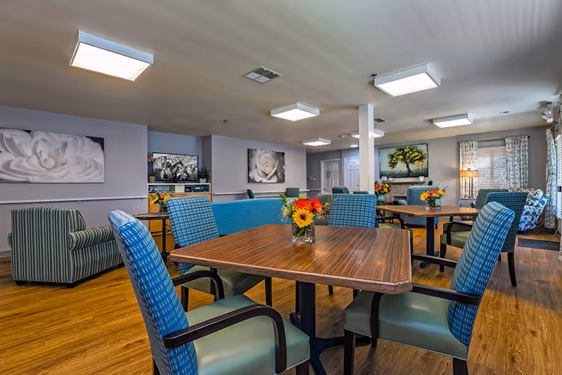 Communal dining and lounge room with wooden tables, upholstered chairs, floral centerpieces, and artwork on the walls.