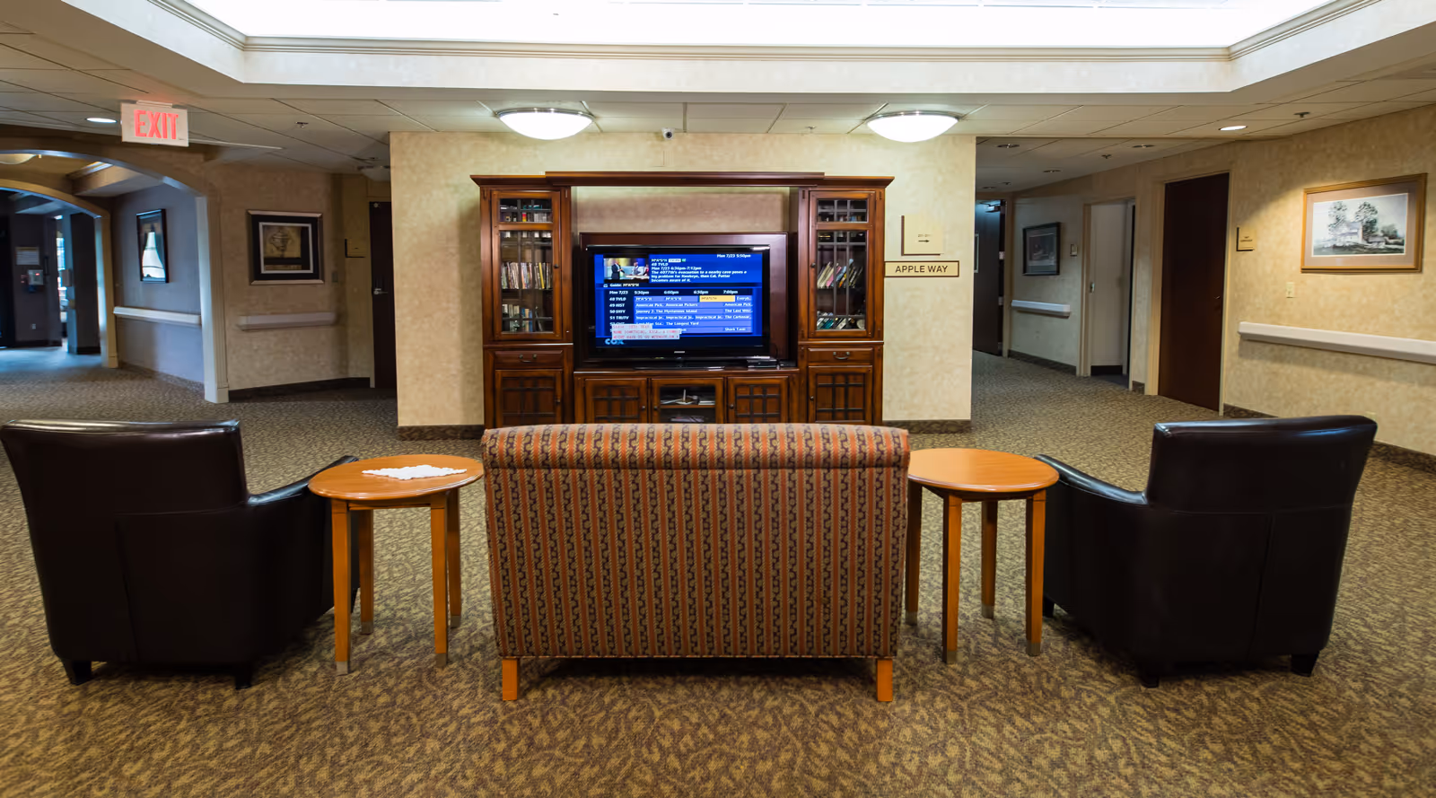 A carpeted common sitting area with two leather chairs, a patterned loveseat and side tables facing a television in a wooden entertainment center in a senior living hallway.