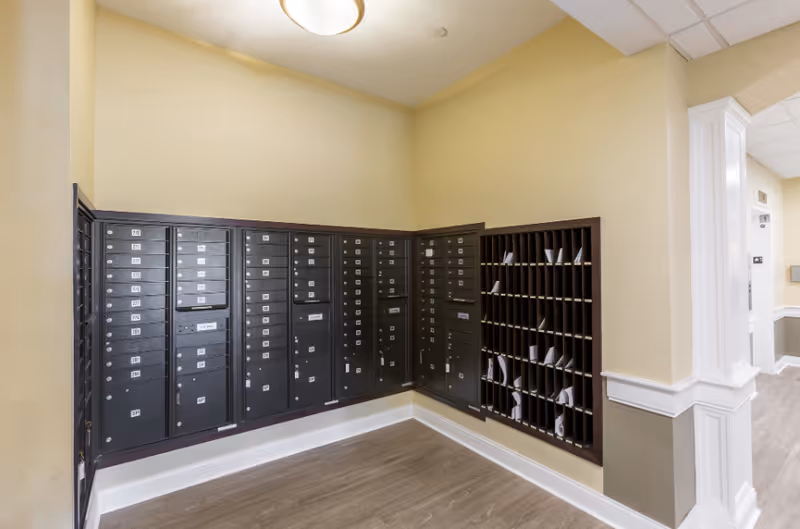 Interior corner of a senior living facility with a wall of black mailboxes and mail slots filled with letters. The walls are painted beige with white trim, and the floor has light wood-style flooring. A ceiling light fixture illuminates the area.