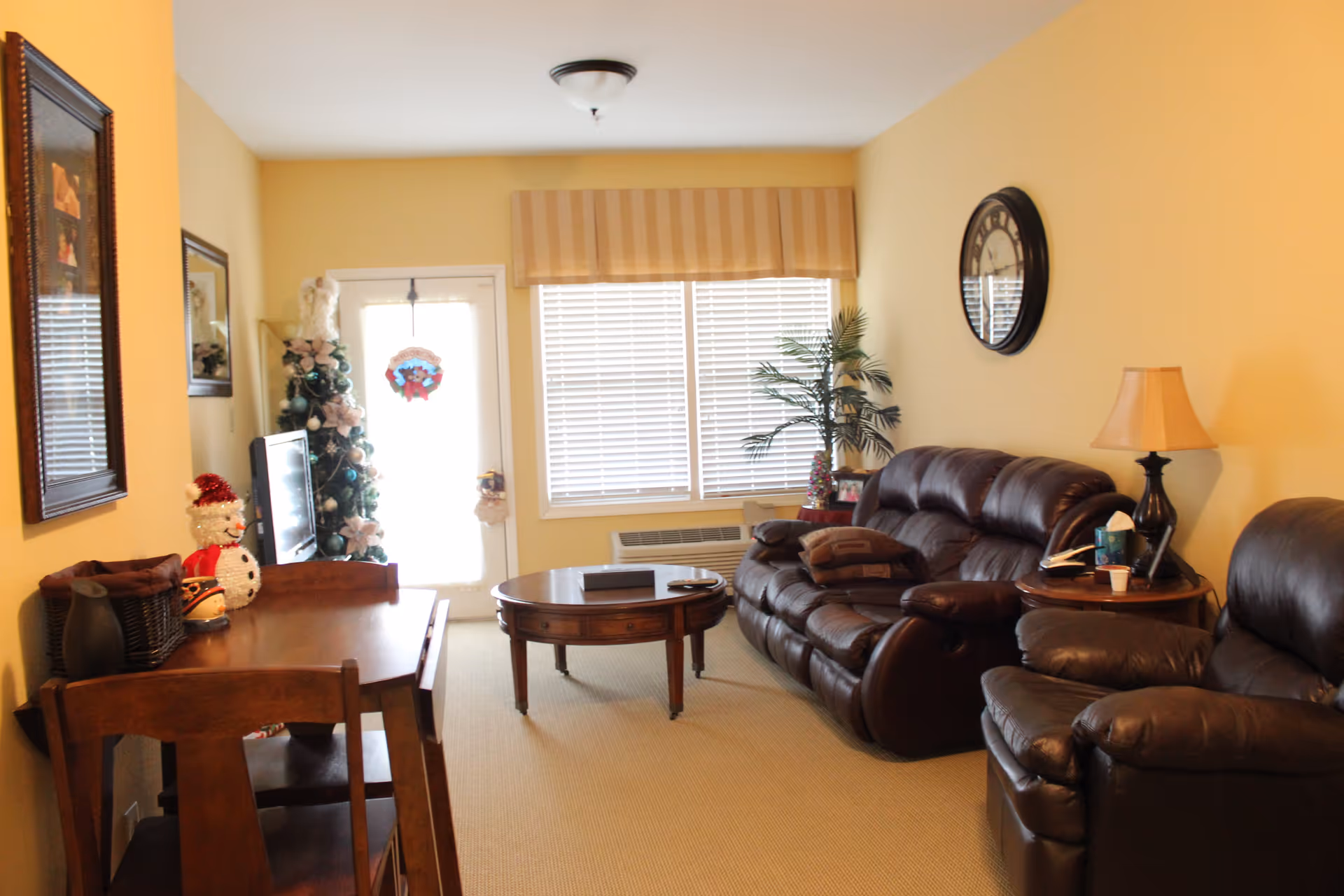 A cozy living room with beige walls and carpet, featuring a dark brown leather sofa and armchair, a round wooden coffee table, a side table with a lamp and tissues, a wall clock, a small artificial palm plant, a dining table with chairs, a Christmas tree, and a door with a holiday wreath.