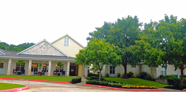 Exterior view of a single-story building with stone and light yellow siding, a covered porch with hanging plants, outdoor seating, and a well-maintained lawn with trees and flower beds.