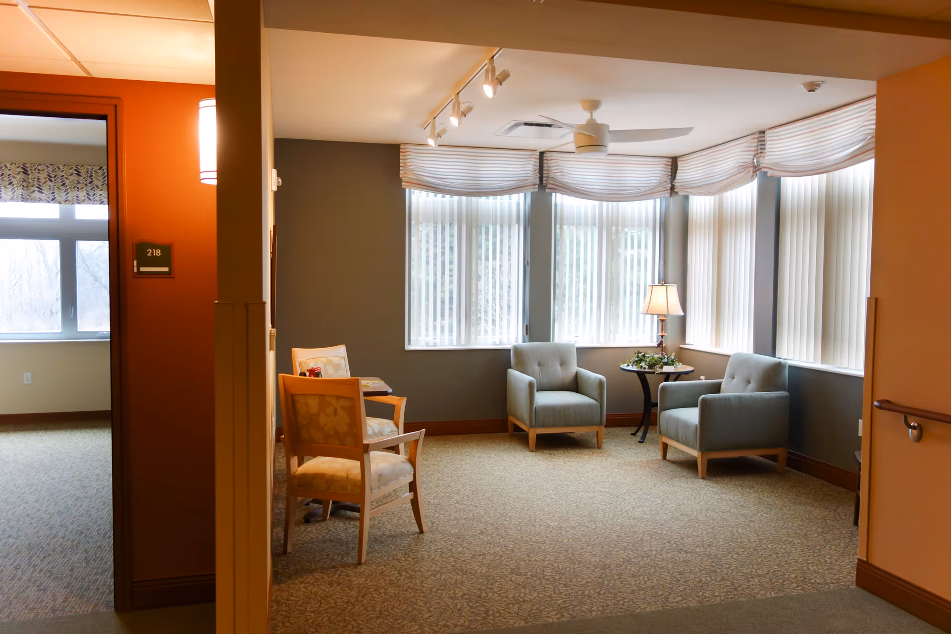 A cozy sitting area in a senior living facility with two light blue armchairs and a small round table with a lamp and decorative greenery. The room has large windows with vertical blinds and striped valances, beige carpet, and soft lighting. To the left, there is a doorway labeled 218 leading to another room with a window.