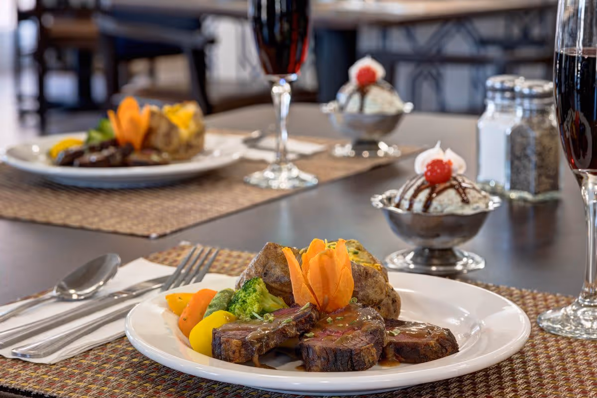 A close-up view of a dining table set with plates of food including sliced steak with sauce, steamed vegetables, and a baked potato garnished with a carved carrot flower. In the background, there are dessert dishes with ice cream topped with chocolate syrup and a cherry, salt and pepper shakers, and glasses of red wine.