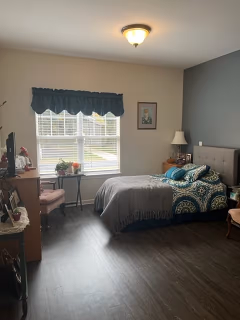 A cozy bedroom with a single bed covered in a patterned blue and white comforter and a gray throw blanket. There is a nightstand with a lamp and picture frames next to the bed. A window with white blinds and a dark blue valance lets in natural light. A small table with plants and a chair are positioned near the window. The room has dark wood flooring and neutral-colored walls, with one accent wall painted dark gray.