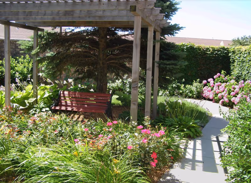 Wooden pergola with a red bench in a landscaped garden surrounded by flowering plants and a paved walkway.