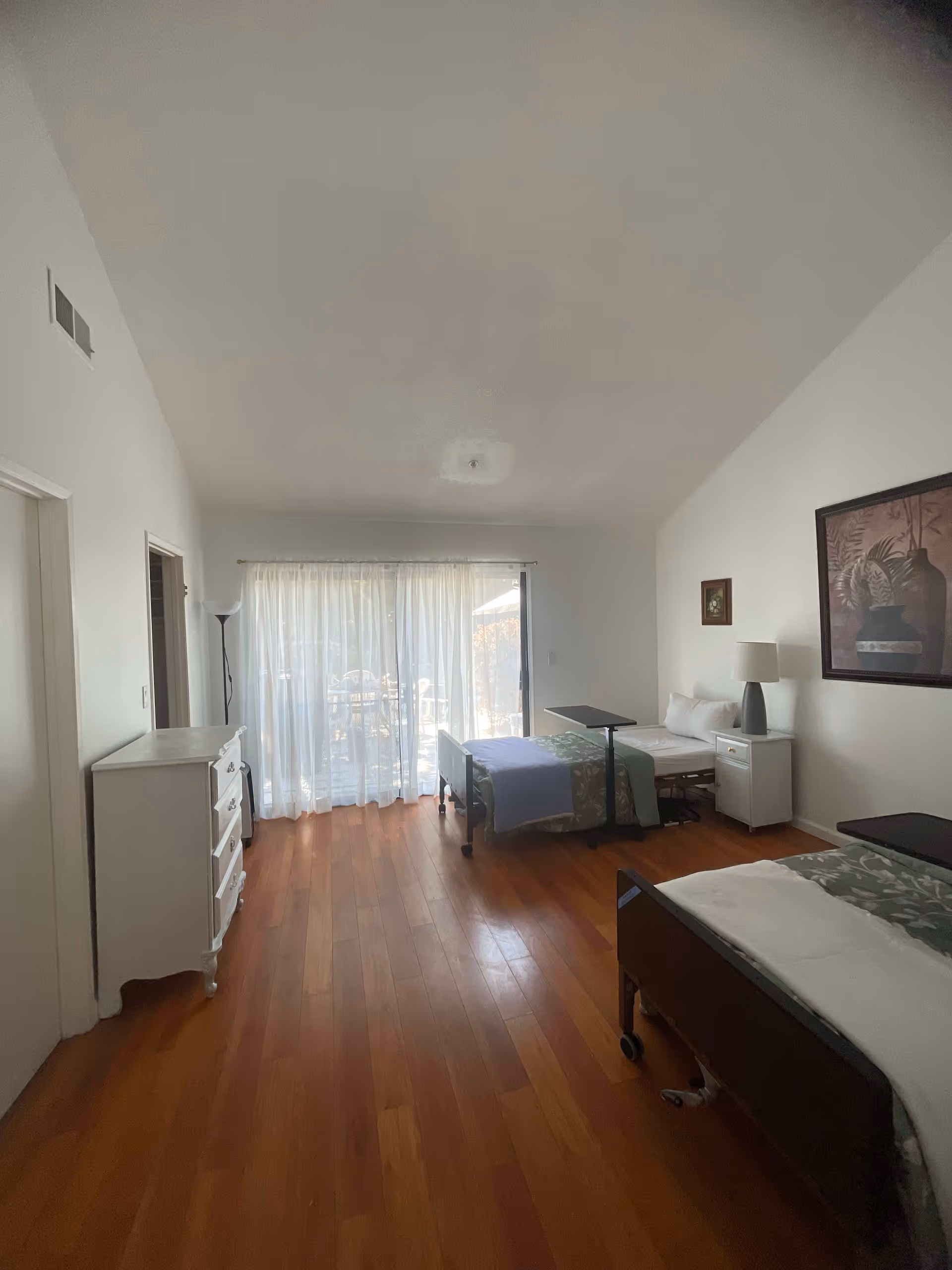 A bright bedroom with two hospital-style beds, each with a green and white blanket and a small overbed table. The room has wooden flooring, white walls, a white dresser on the left, a nightstand with a lamp between the beds, and a large window with sheer white curtains letting in natural light. There is a framed picture on the right wall.