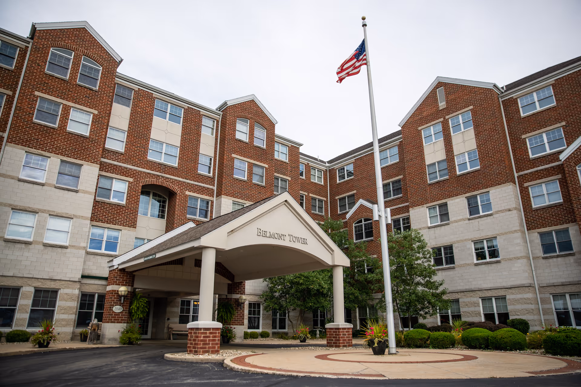 Front entrance of a multi-story brick senior living building labeled 'Belmont Tower' with a covered portico and an American flag.