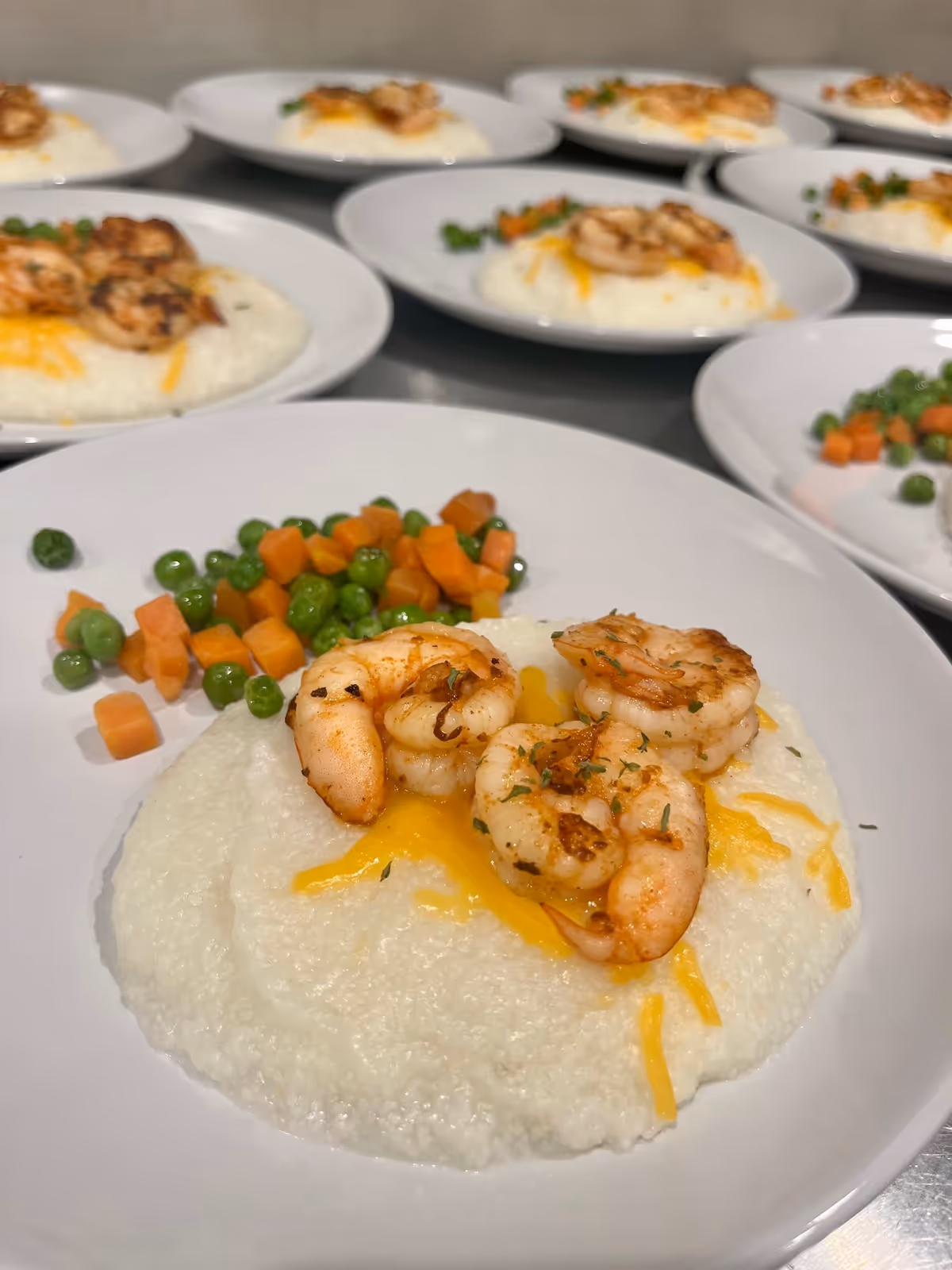 Plates of cooked shrimp served on a bed of creamy grits with melted cheese, accompanied by a side of diced carrots and peas, arranged on a table in a kitchen or dining area.