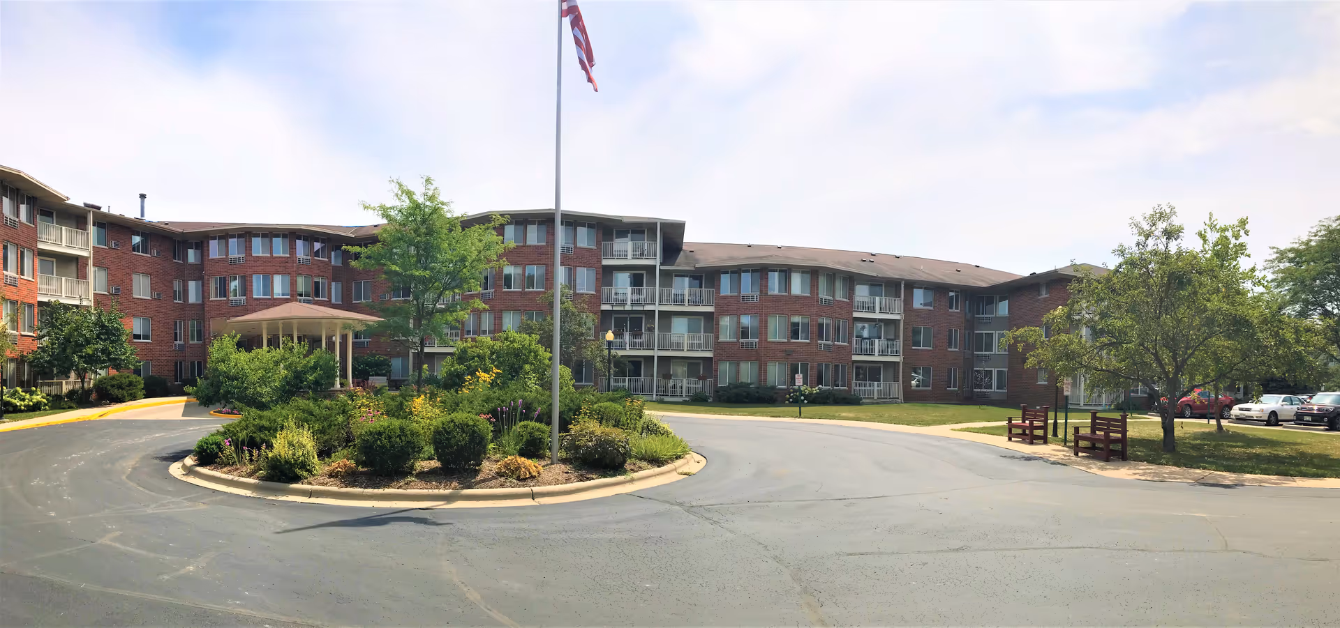 Exterior view of a multi-story senior living facility with red brick walls and multiple windows. There is a circular driveway with a landscaped island in the center featuring bushes and a tree. An American flag is flying on a flagpole in the middle of the island. Several cars are parked on the right side near trees and benches.