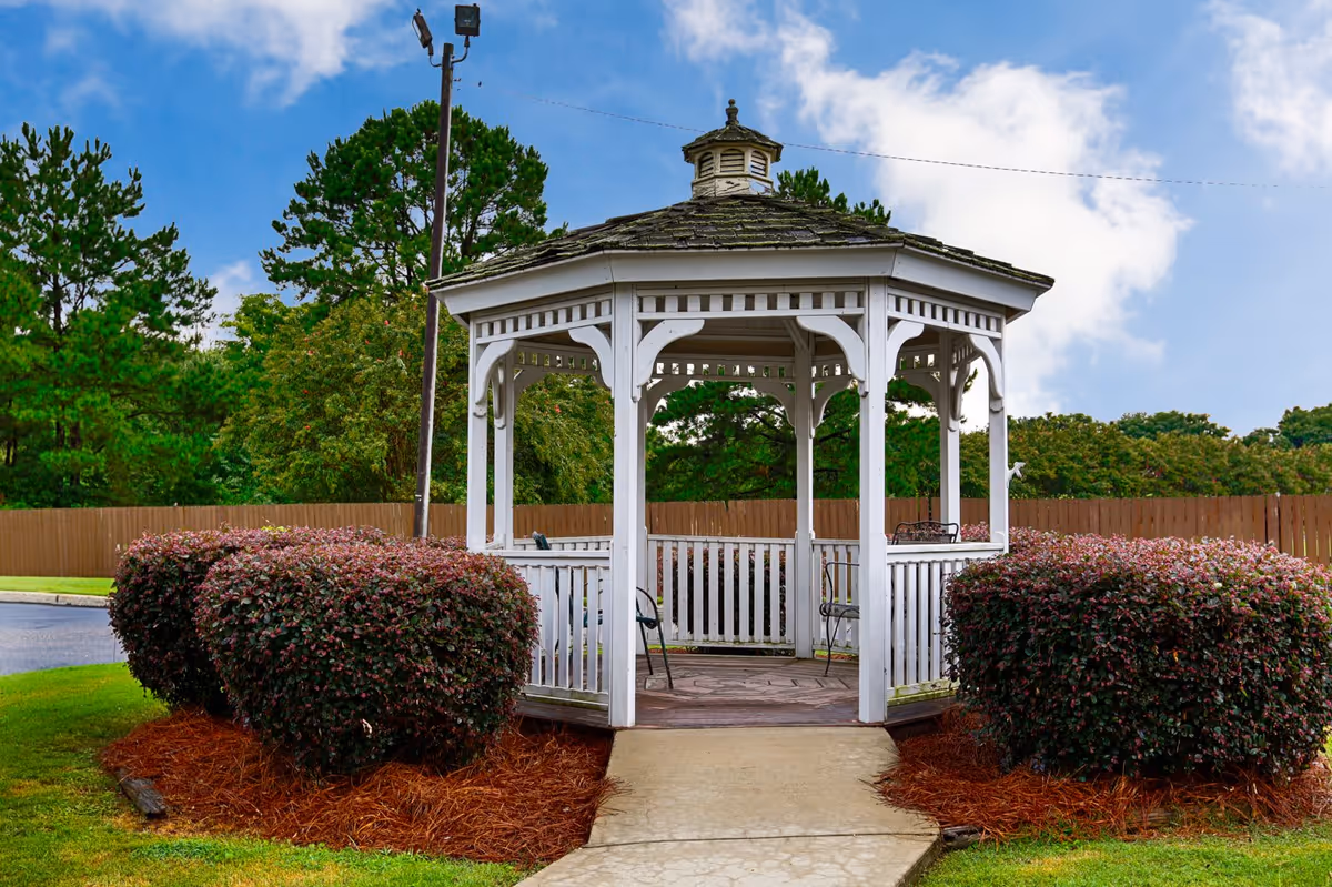 A white wooden gazebo with a shingled roof situated in a landscaped outdoor area with trimmed bushes and pine trees in the background under a partly cloudy blue sky.