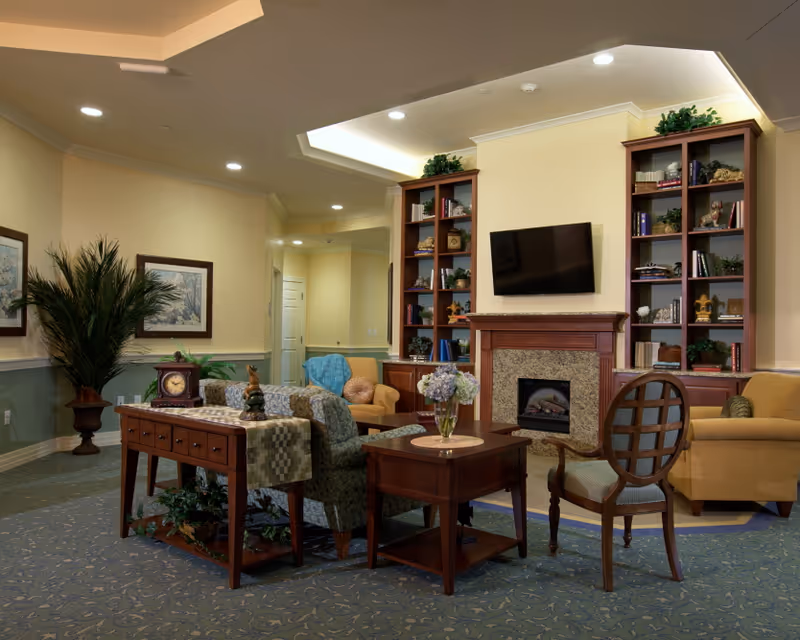 Cozy furnished common living area with sofas, chairs, a coffee table, fireplace, wall-mounted TV, and built-in bookshelves.