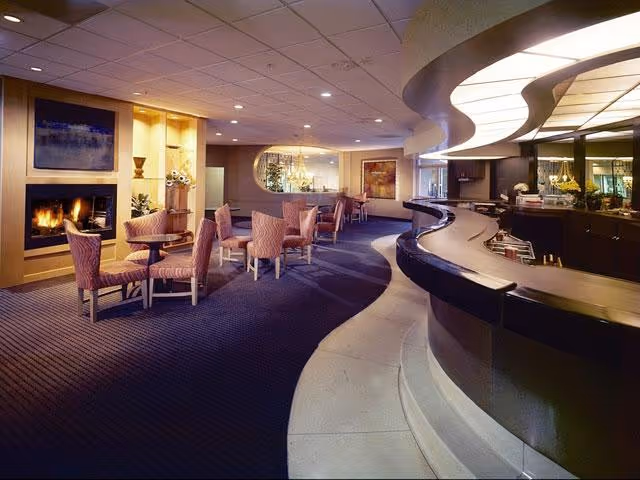 Interior view of a senior living facility lounge area with a curved reception desk on the right, several small tables with upholstered chairs, a lit fireplace on the left, and decorative artwork on the walls.