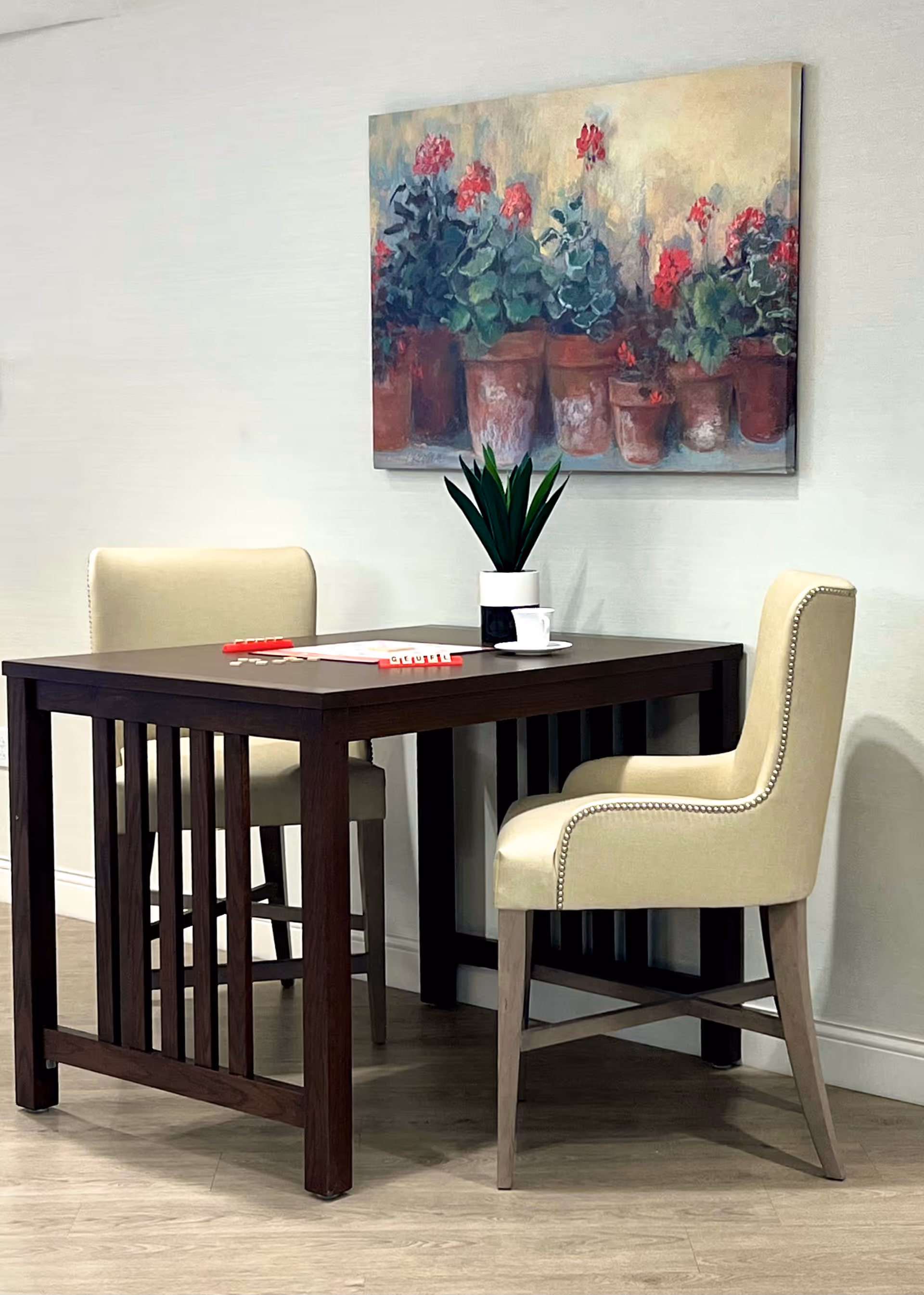 A small dark wooden table with two beige upholstered chairs on either side. On the table, there is a small potted plant, a white cup and saucer, and a Scrabble game with tiles placed on the board. A painting of potted red flowers hangs on the light-colored wall behind the table.
