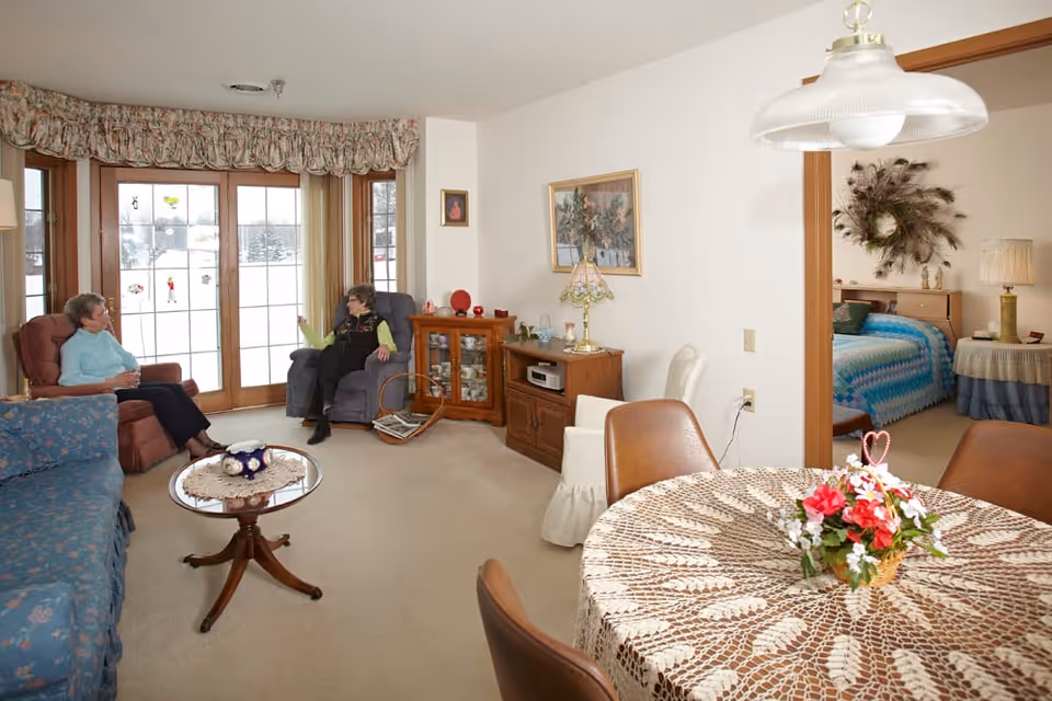 A cozy living room area with two elderly women sitting and conversing on armchairs near large windows with floral valances. The room features a blue floral sofa, a round wooden coffee table with a doily and decorative items, a wooden cabinet with a lamp, and a dining table with a lace tablecloth and a flower centerpiece. A bedroom with a blue bedspread and a wreath on the wall is visible through an open doorway.