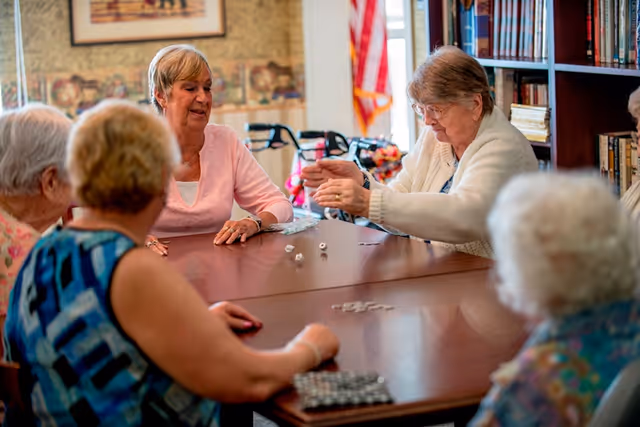 A group of elderly women sitting around a wooden table playing a game with dice and tiles in a cozy room with bookshelves and a window with curtains.