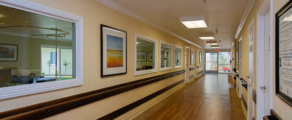 Well-lit interior hallway in a senior living facility with handrails, framed artwork, and windows into adjacent rooms.
