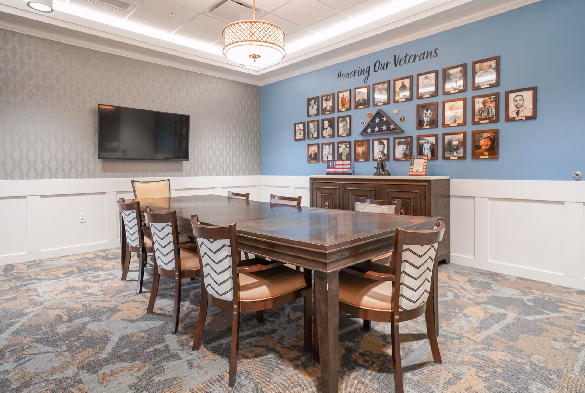 A communal dining/meeting room with a large wooden table and chairs, a wall-mounted TV, and a display honoring veterans.