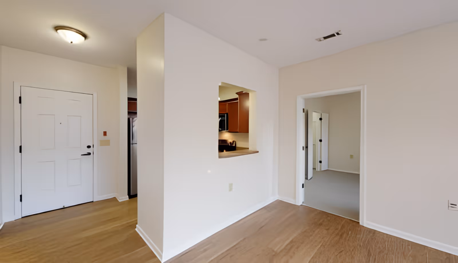 Interior view of an apartment or living space showing a white front door on the left, a small pass-through window opening into a kitchen with wooden cabinets, and an open doorway leading to another room with carpeted flooring. The walls are painted white and the floor in the main area is wood.