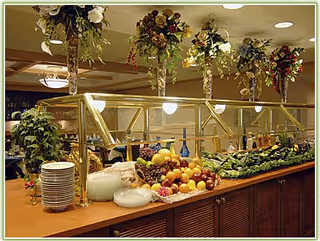 Buffet-style dining area with a variety of fresh fruits and vegetables displayed on a wooden counter with a glass sneeze guard. Plates are stacked on one side, and decorative hanging flower arrangements are above the counter. Warm lighting illuminates the space.