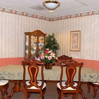 A dining room with a large wooden dining table covered with a green patterned tablecloth. Six ornate wooden chairs with cushioned seats surround the table. A floral centerpiece is placed in the middle of the table. In the background, there is a wooden china cabinet with glass doors displaying dishes, a framed artwork on the wall, and a potted plant. The walls have wallpaper with a decorative border near the ceiling, and the ceiling has a light fixture.