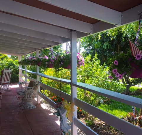 Covered porch with white railings, hanging flowers, and chairs overlooking a sunny garden.
