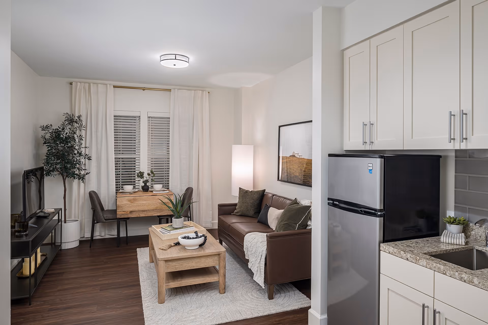 A cozy living room area with a brown leather sofa adorned with pillows and a throw blanket, a wooden coffee table with decorative items, a small dining table with two chairs set near a window with white curtains, a flat-screen TV on a black stand, and a compact kitchen area with a stainless steel mini refrigerator and granite countertop.