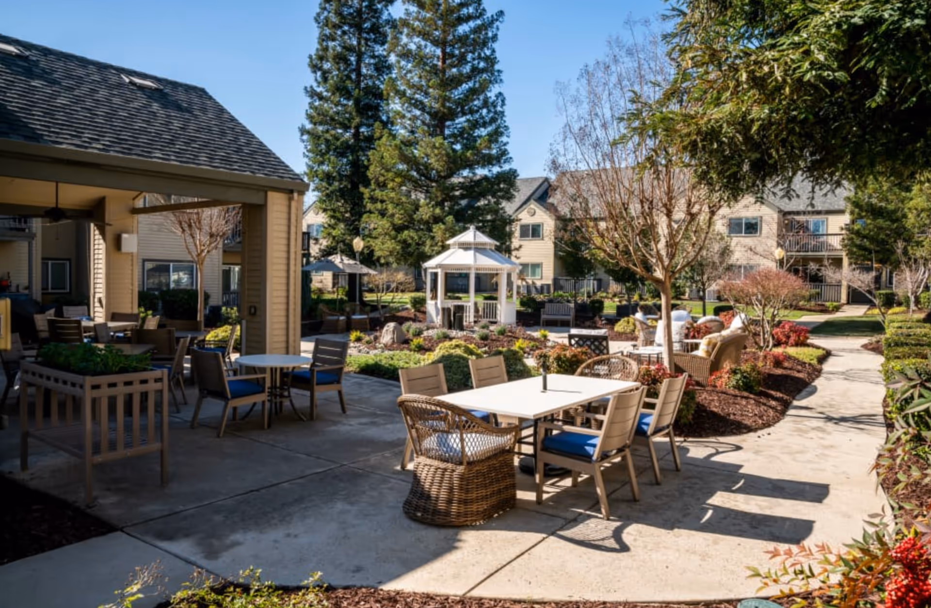 Sunlit courtyard of a senior living community with patio tables and chairs, a gazebo, landscaped beds, and surrounding residential buildings.