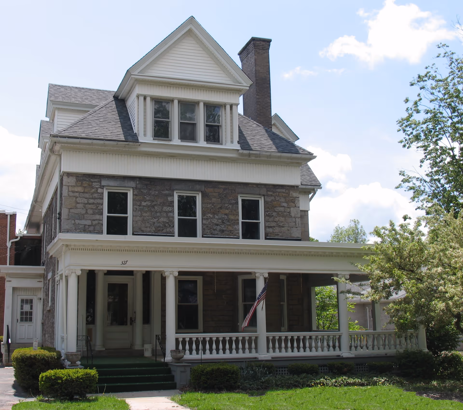 A large, three-story stone and wood house with a covered front porch supported by white columns. The house has multiple windows, a chimney, and an American flag hanging on the porch. There is green grass and bushes in front of the house under a partly cloudy sky.