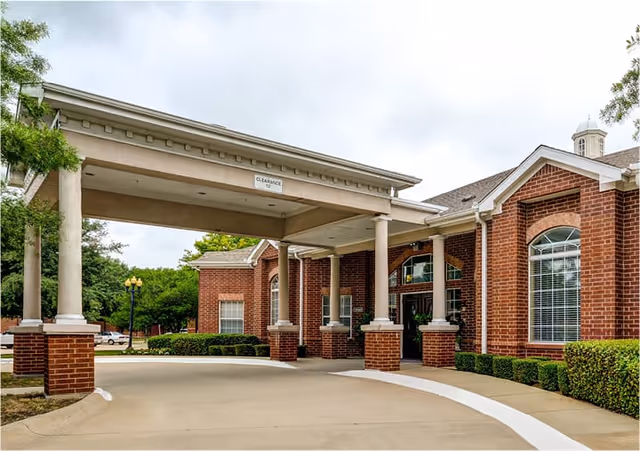 Exterior view of a senior living facility with a covered entrance supported by columns, red brick walls, large windows with white blinds, and neatly trimmed bushes along the walkway.