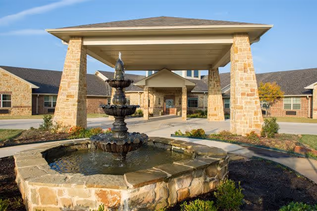 Stone fountain with three tiers in front of a covered entrance supported by stone pillars at a single-story brick building under a clear blue sky.