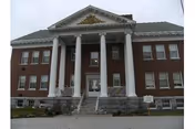 Front exterior view of a large brick building with white columns and a triangular pediment above the entrance, featuring steps leading up to the main door.