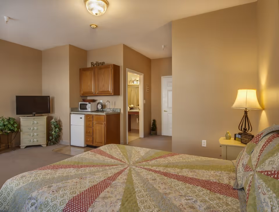 A cozy senior living bedroom with a patterned quilt on the bed, a nightstand with a lamp and decorative items, a small kitchenette area with wooden cabinets, a microwave, mini fridge, and a sink. There is a TV on a small dresser, some plants, and a doorway leading to a bathroom.