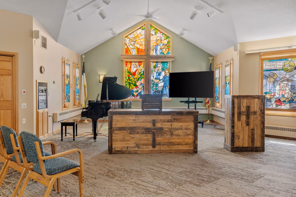 Interior of a chapel or worship room with stained glass windows depicting religious scenes, a wooden altar with a cross, a grand piano, a TV screen on a stand, and several chairs arranged in rows.