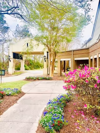 Outdoor pathway leading to the entrance of a brick building with a covered driveway. The scene includes blooming flowers and green trees under a bright sky.