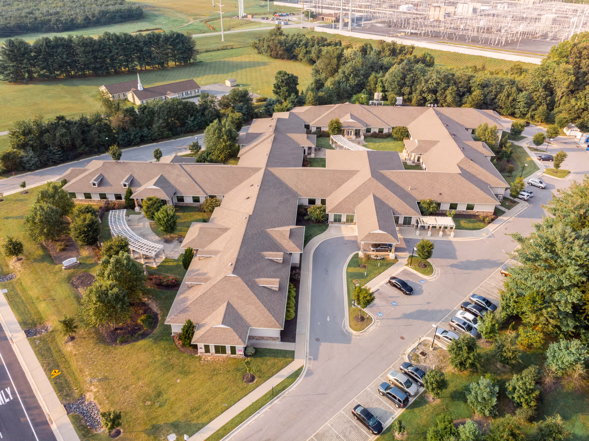 Aerial view of a single-story senior living facility complex with connected beige buildings, a circular drive and parking lot, and surrounding lawns and trees.