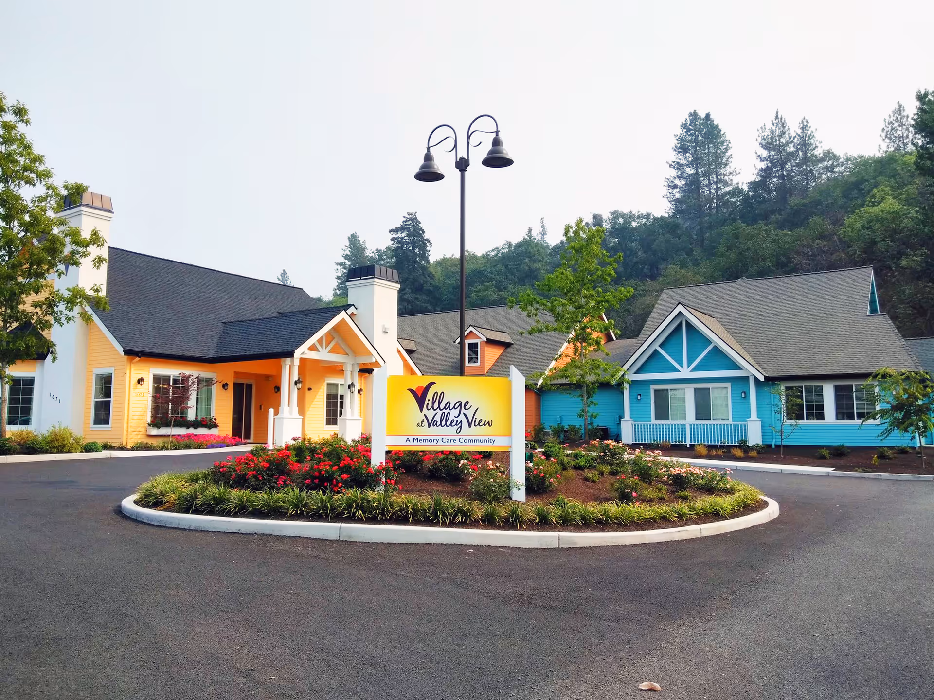 Exterior view of Village at Valley View, a memory care community, showing a circular driveway with landscaped flower beds in the center. The building has a welcoming appearance with yellow and blue painted sections, white trim, and a covered entrance. Trees and greenery surround the facility.