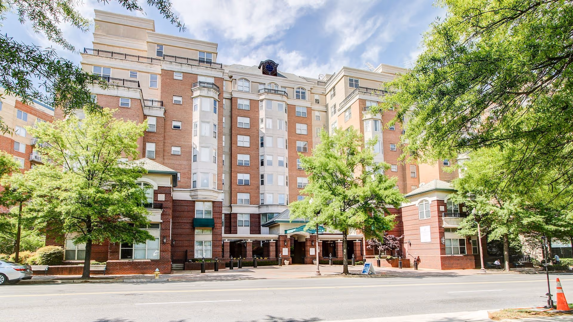 Exterior view of a multi-story senior living facility building named Vitality Living Arlington, featuring a brick and beige facade with multiple windows, trees lining the street in front, and a clear blue sky with some clouds above.