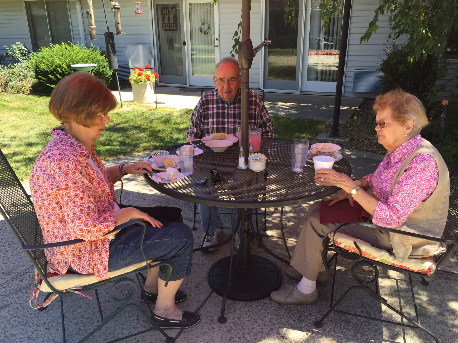 Three elderly people sitting around a round metal outdoor table with an umbrella pole in the center, enjoying drinks and snacks on a sunny day in a garden patio area outside a building.