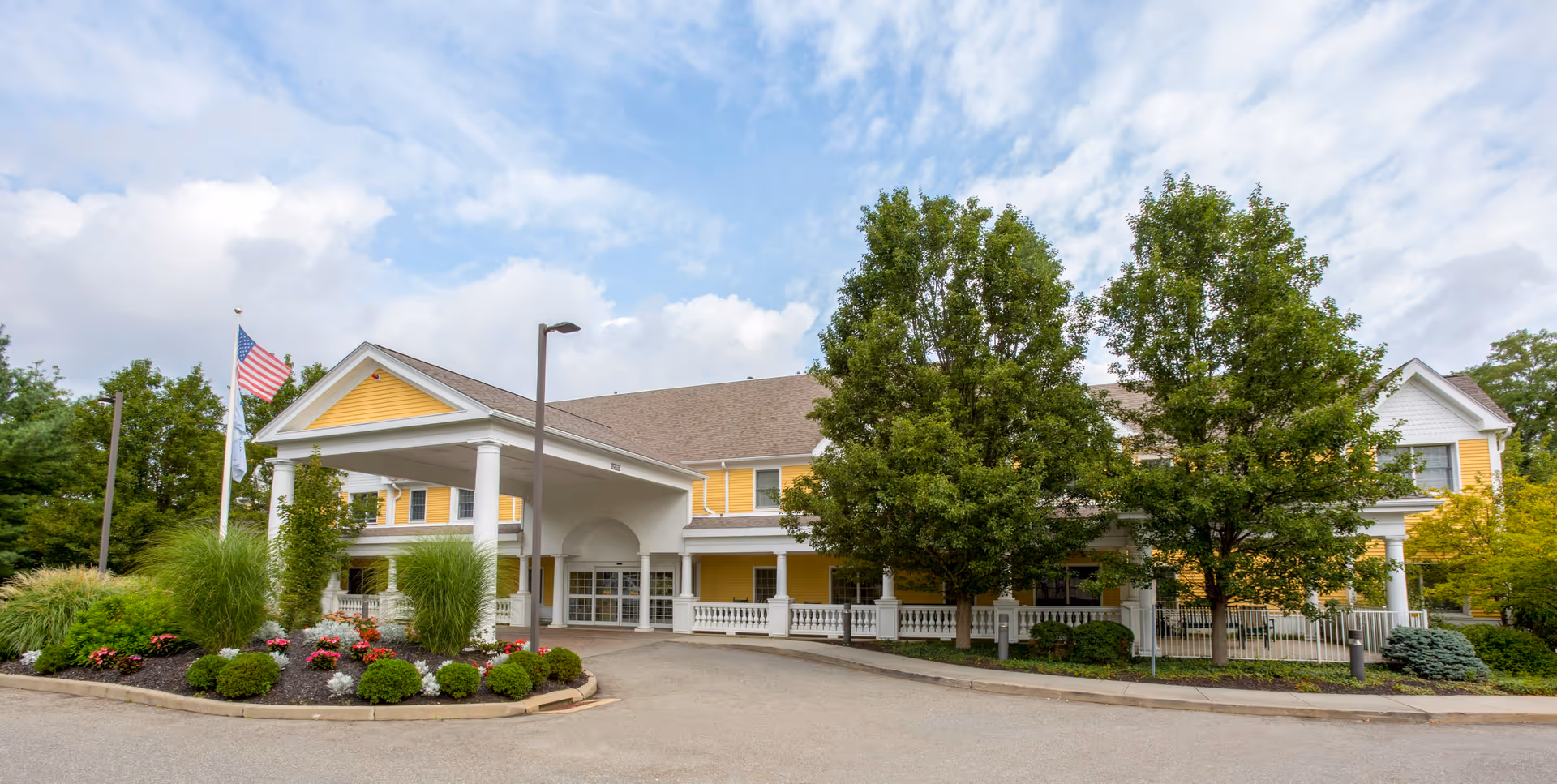 Exterior view of a senior living facility building with a covered entrance, yellow siding, white trim, and a landscaped area with bushes and flowers. Two flags, including an American flag, are visible near the entrance under a partly cloudy sky.