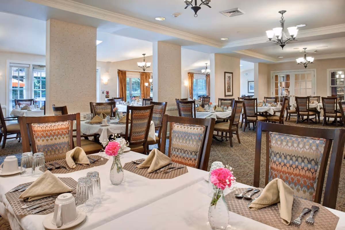 Bright dining room with multiple set tables, wooden chairs, chandeliers, and small floral centerpieces.