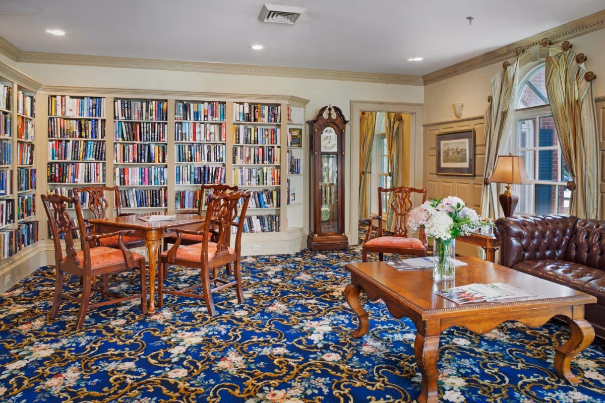 A cozy and elegant common room with a blue floral carpet, wooden furniture including a table with four chairs, a coffee table with a vase of flowers, a leather tufted sofa, and a grandfather clock. The room has built-in bookshelves filled with books, large windows with striped curtains, and warm lighting.