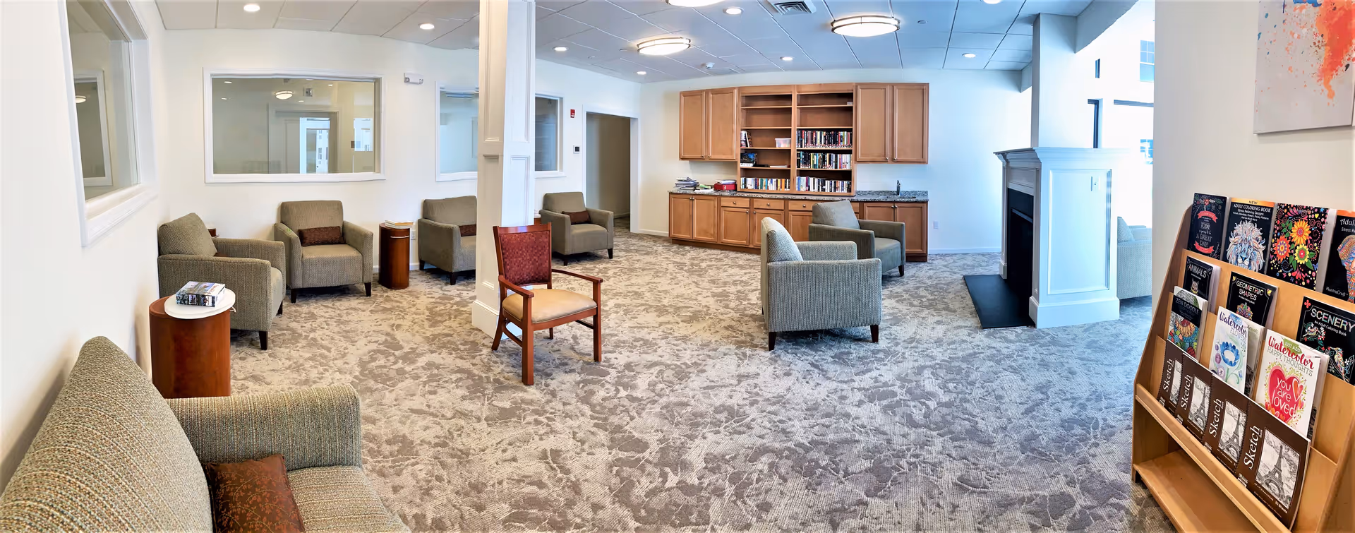 A spacious senior living common area with multiple armchairs and a single wooden chair arranged around a central column. There is a built-in wooden bookshelf and cabinet unit against the far wall, filled with books and other items. The room has a patterned carpet, white walls, ceiling lights, and a fireplace on the right side. A wooden rack with various coloring books and sketch pads is visible in the foreground on the right.