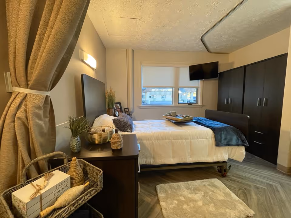Well-appointed senior bedroom with a made single bed, bedside table and dresser, a wall-mounted TV, window, and decorative baskets.