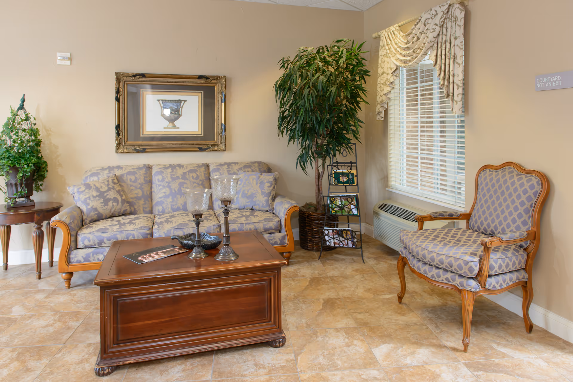A cozy living room area with a patterned sofa and matching armchair, a wooden coffee table with decorative candle holders, a side table with a plant, a tall potted plant, a window with blinds and a decorative valance, and a framed picture on the wall.
