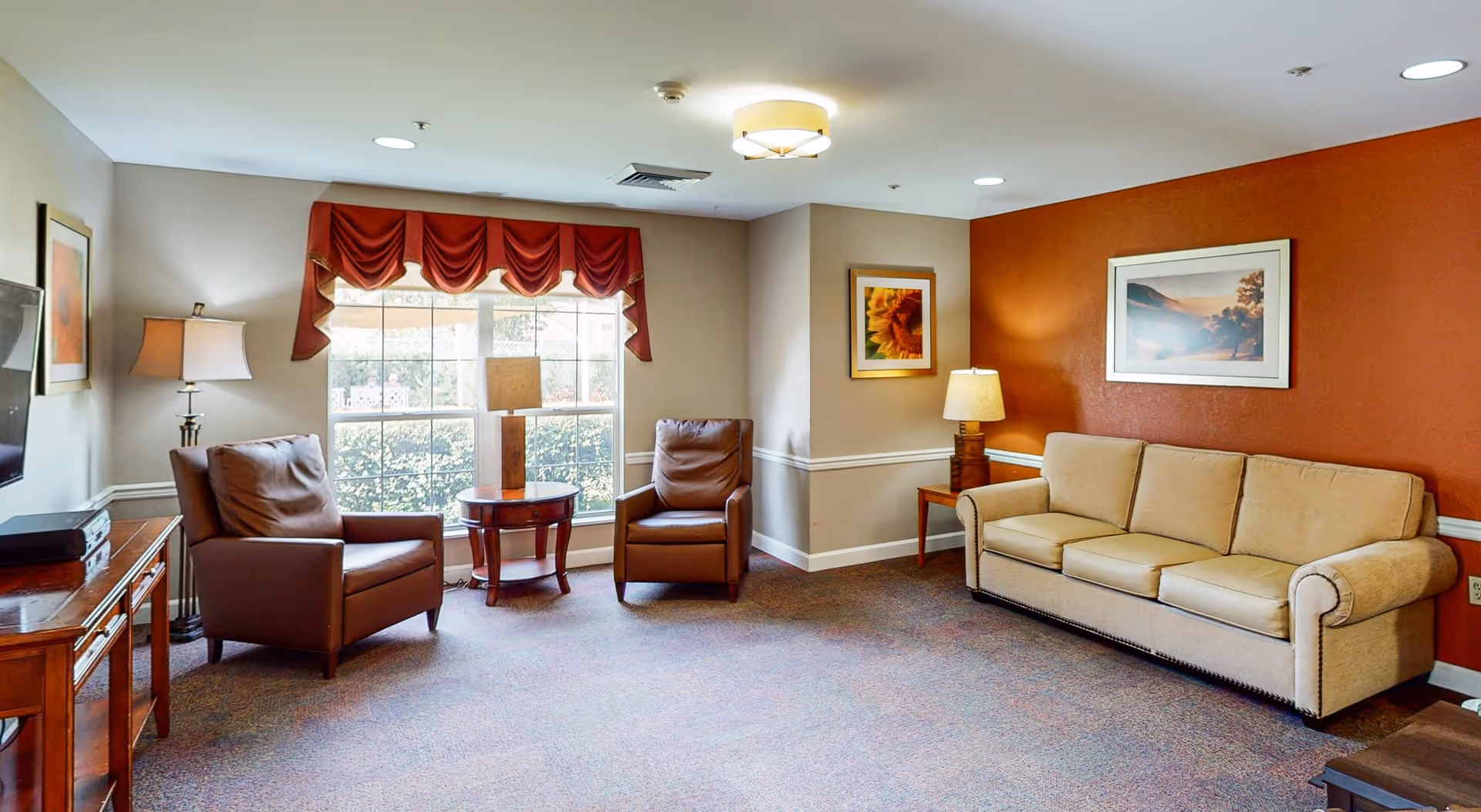 A cozy living room with a beige couch against a burnt orange accent wall, two brown leather armchairs near a large window with red valance curtains, a wooden side table with a lamp, and framed artwork on the walls.