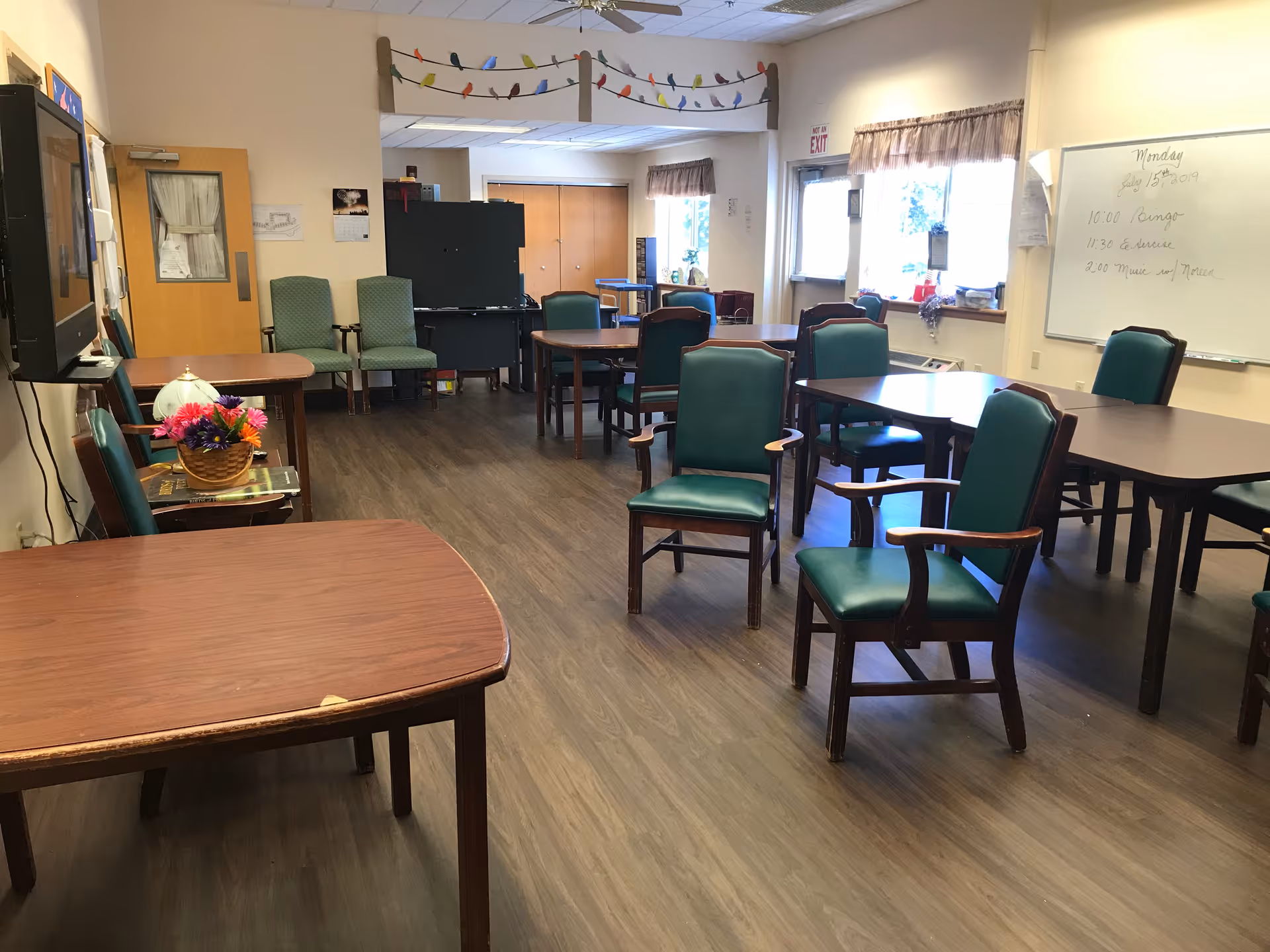 Community activity/dining room with multiple tables and green upholstered chairs, a whiteboard, TV, and windows.