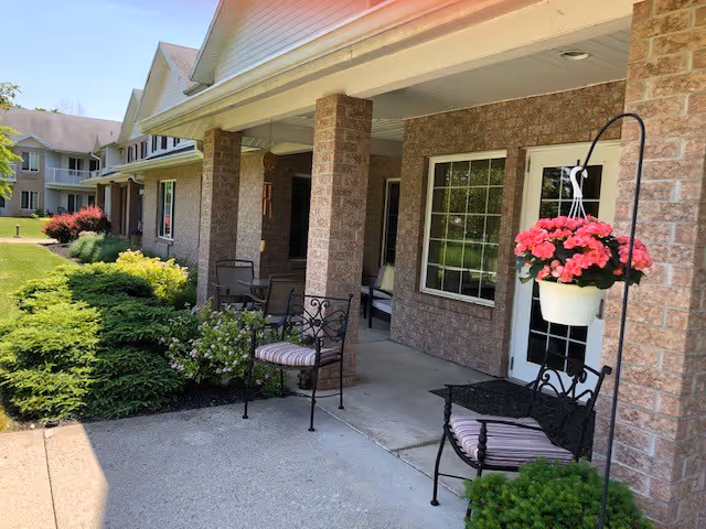 Covered front porch of a senior living building with brick columns, outdoor chairs, hanging potted flowers, and landscaped greenery.