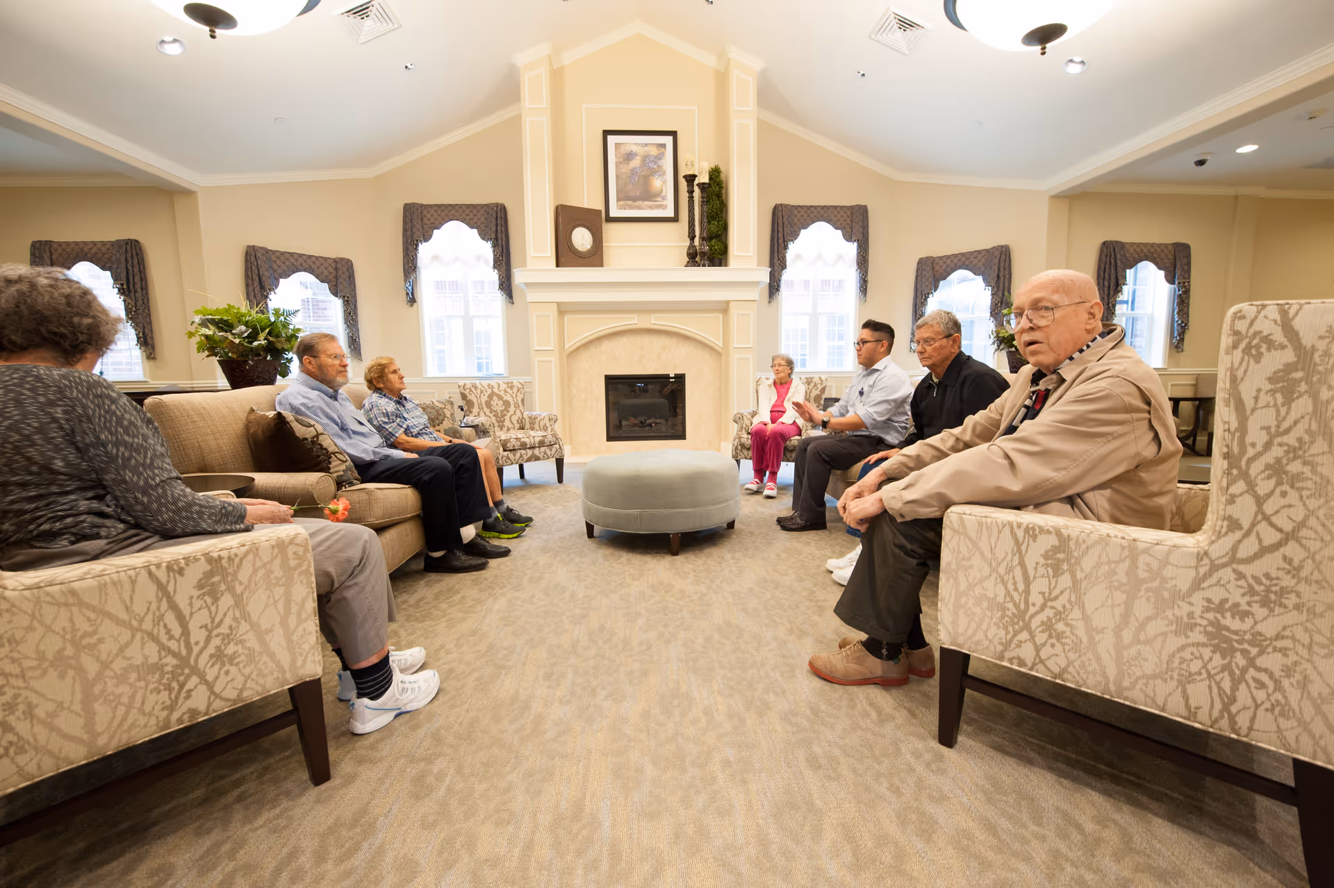 A group of elderly people sitting in a spacious, well-lit living room with beige walls, patterned armchairs, a central round ottoman, and a fireplace with decorative items above it. The room has multiple windows with valances and a carpeted floor.