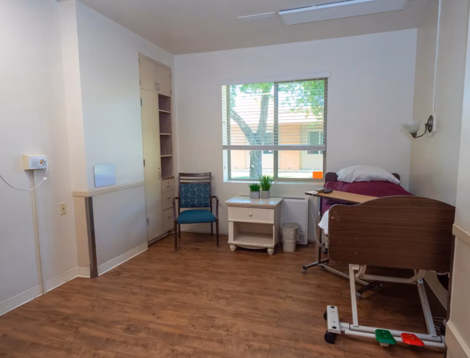 A simple, clean room with a hospital-style bed with a purple blanket and white pillow, a small white nightstand with two small potted plants, a blue cushioned chair, a window with blinds showing a tree outside, and wooden flooring.