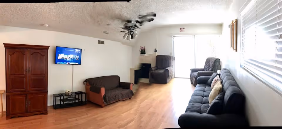 Spacious common living room with sofas and armchairs, a wall-mounted TV, wooden cabinet, ceiling fan, and sliding glass exit door.